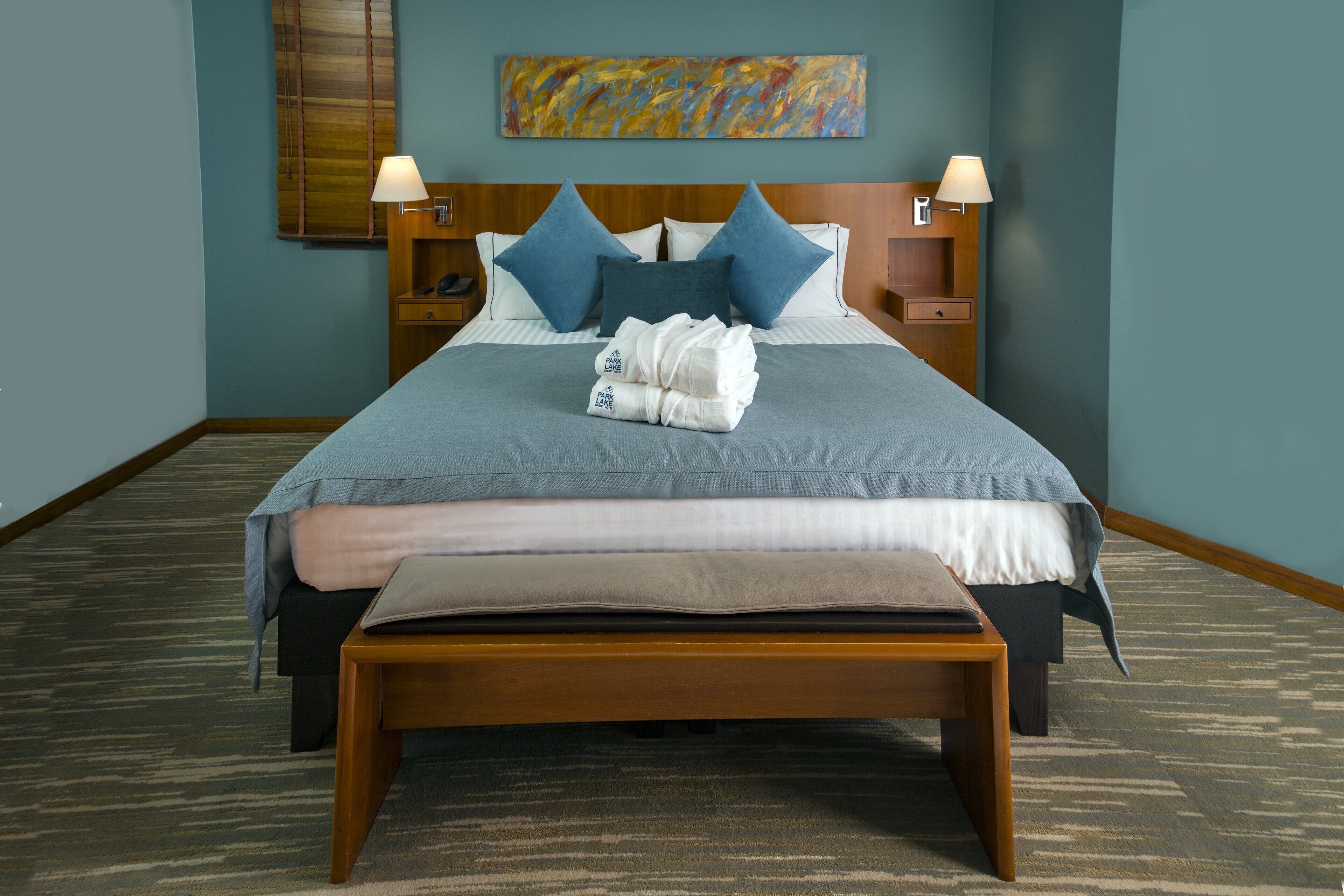A neatly made bed with a blue bedspread, two pillows, and a folded towel. There is a wooden headboard, bedside tables with lamps, and a wall-mounted painting.