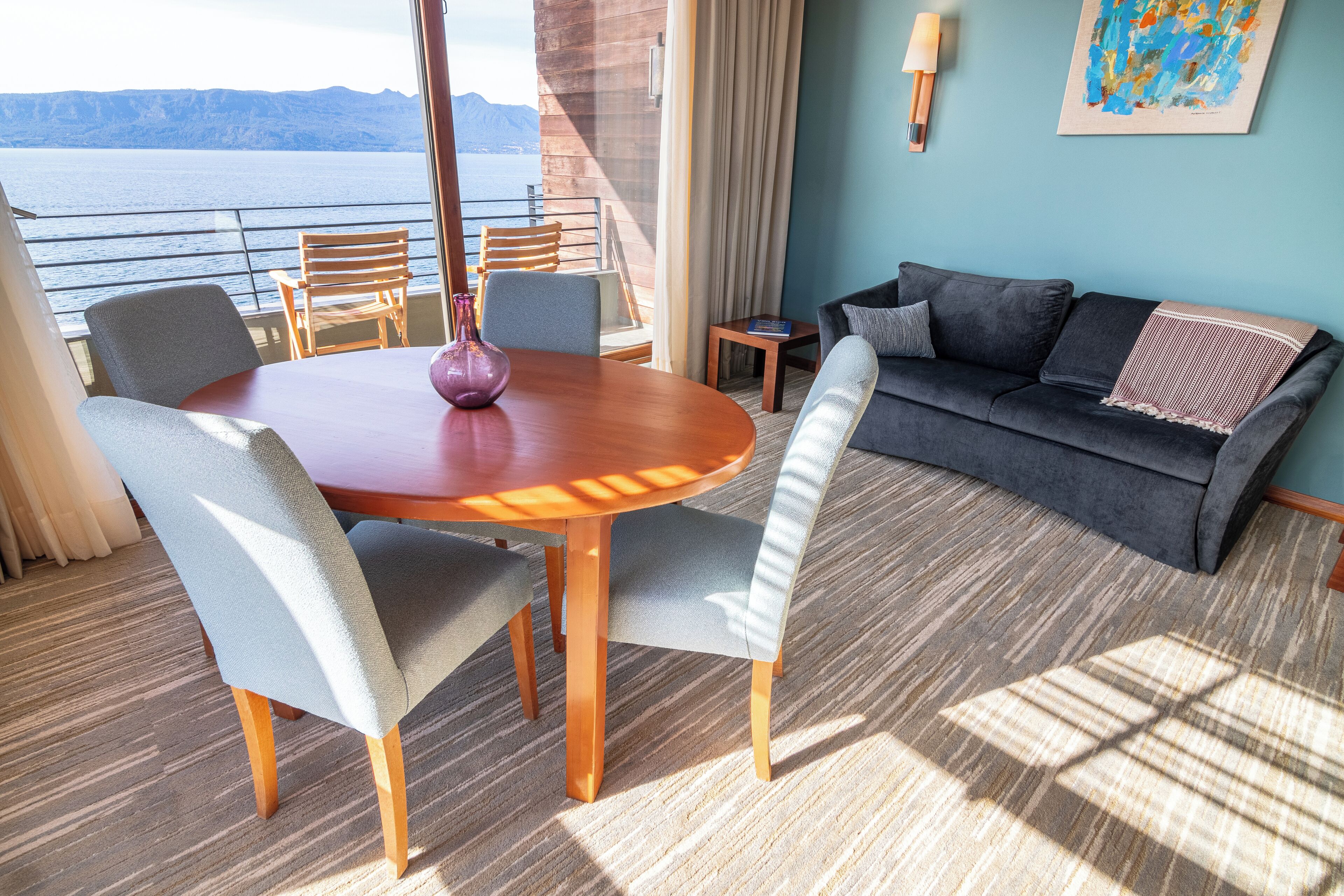 A room with a wooden dining table, grey upholstered chairs, a dark grey sofa, and a view of the sea.