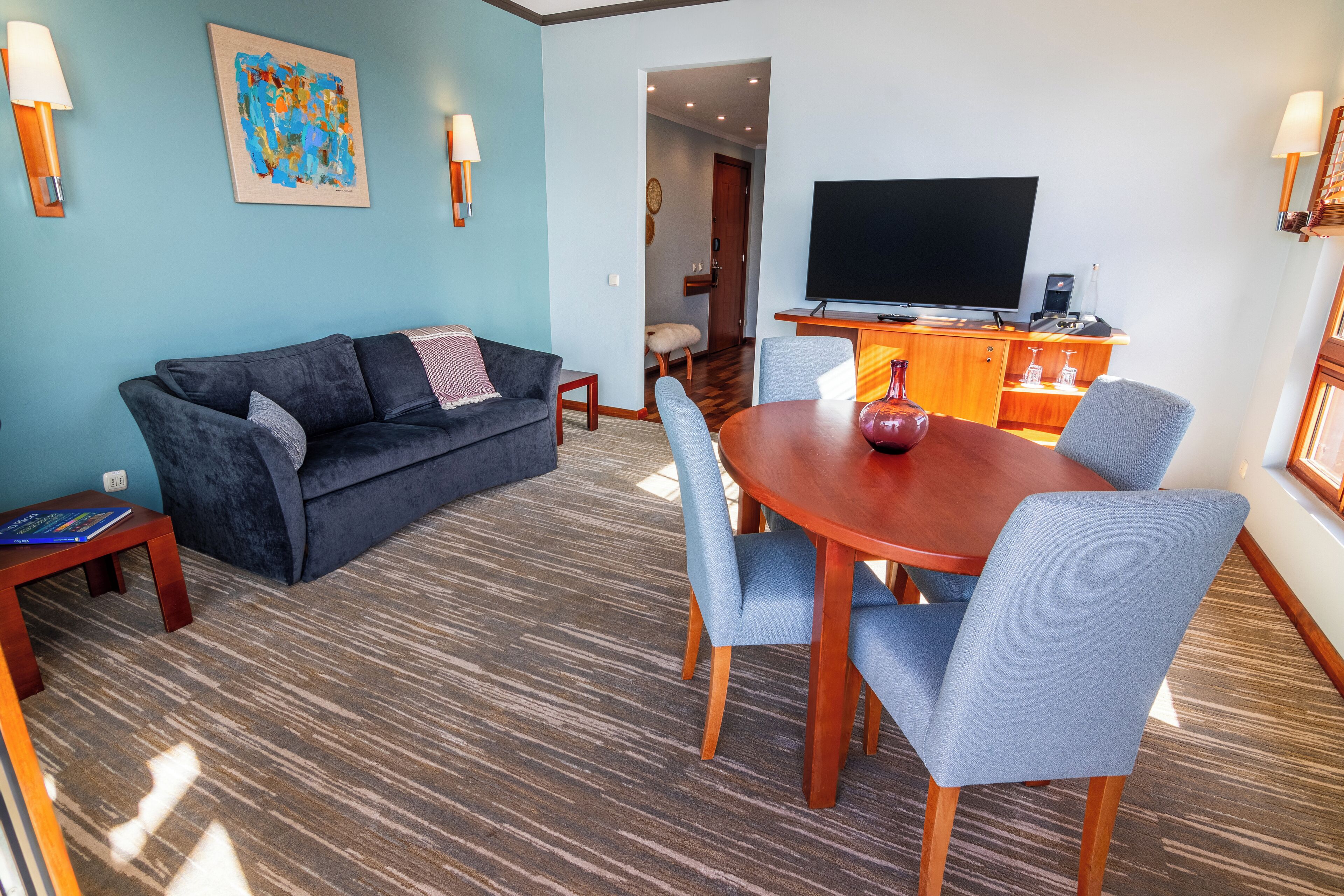 A living room with a dark blue sofa, a wooden dining table with blue chairs, a flat-screen TV, and a small side table.