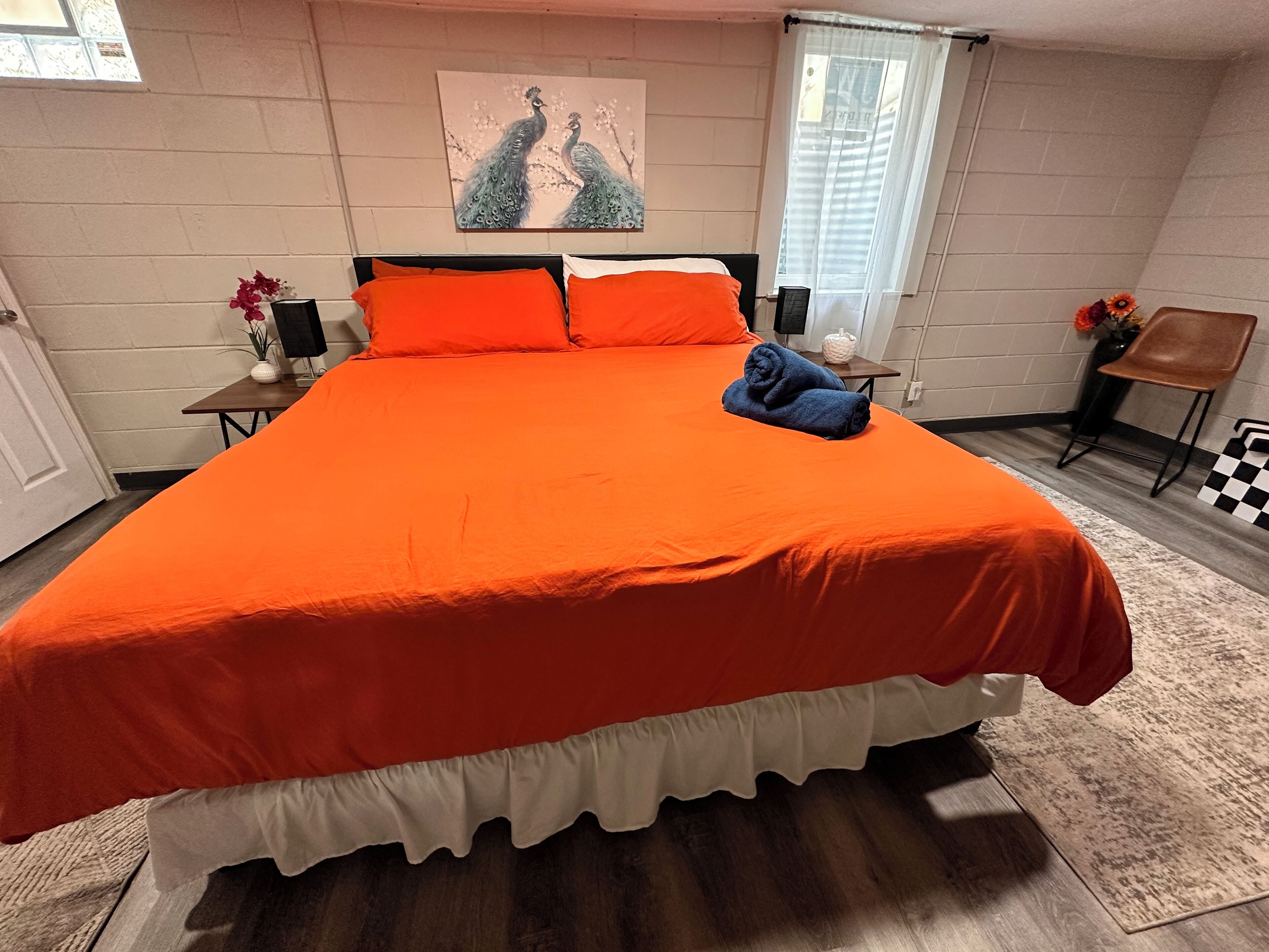 A bedroom with a large bed, a window with blinds, a painting on the wall, and a small table with a vase of flowers.