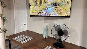 Een woonkamer met een aan de muur bevestigde televisie waarop een natuurscène wordt vertoond, een kleine ventilator en een decoratief olifantje op een houten tafel.