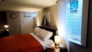 Een slaapkamer met een bed, nachtkastje, lamp, raam met zonwering, wandklok en een deur.
