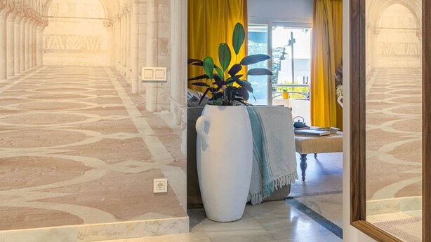 A hallway with a patterned floor leading to a room with a large plant, yellow curtains, and a view of the outside. - Puerto Banús Cozy and Luxe Home (Marbella)