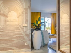 Un pasillo con piso estampado que lleva a una habitación con una planta grande, cortinas amarillas y vistas al exterior.