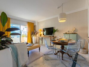 Una sala de estar moderna con comedor, un ventanal con cortinas amarillas, un televisor de pantalla plana y una planta en maceta.