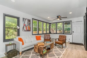Un salon spacieux avec un canapé blanc, deux fauteuils en cuir, une table basse en bois et de grandes fenêtres donnant sur l’extérieur.