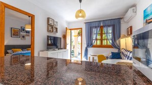 Een moderne woonkamer met een groot granieten eiland, een flatscreen televisie, een zithoek met een gele stoel en uitzicht op het buitenleven door een raam met blauwe gordijnen.