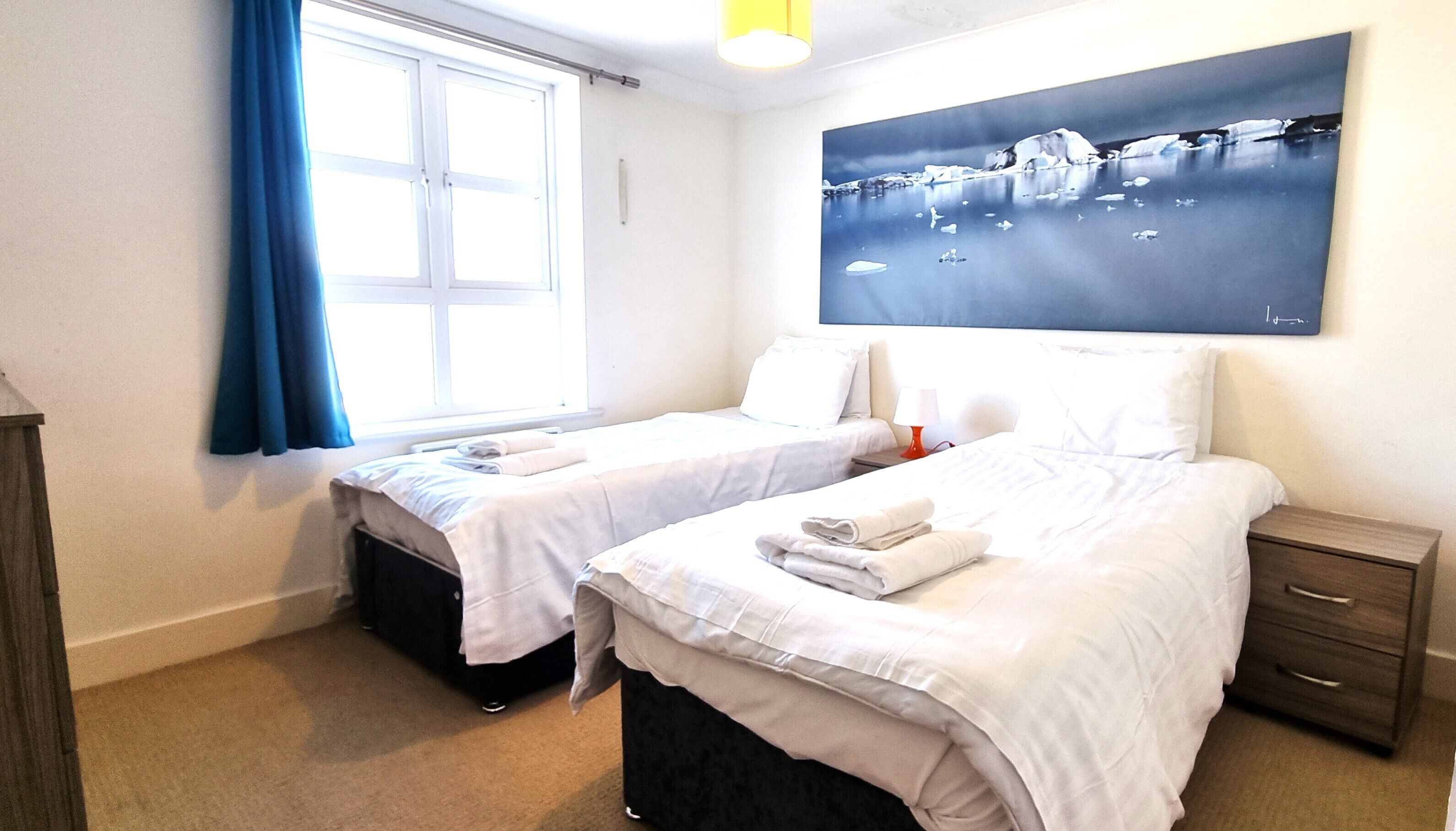 Ein Hotelzimmer mit zwei Einzelbetten, einem Nachttisch mit Lampe, einem Fenster mit blauen Vorhängen und einem Wandbild von Eisbergen.