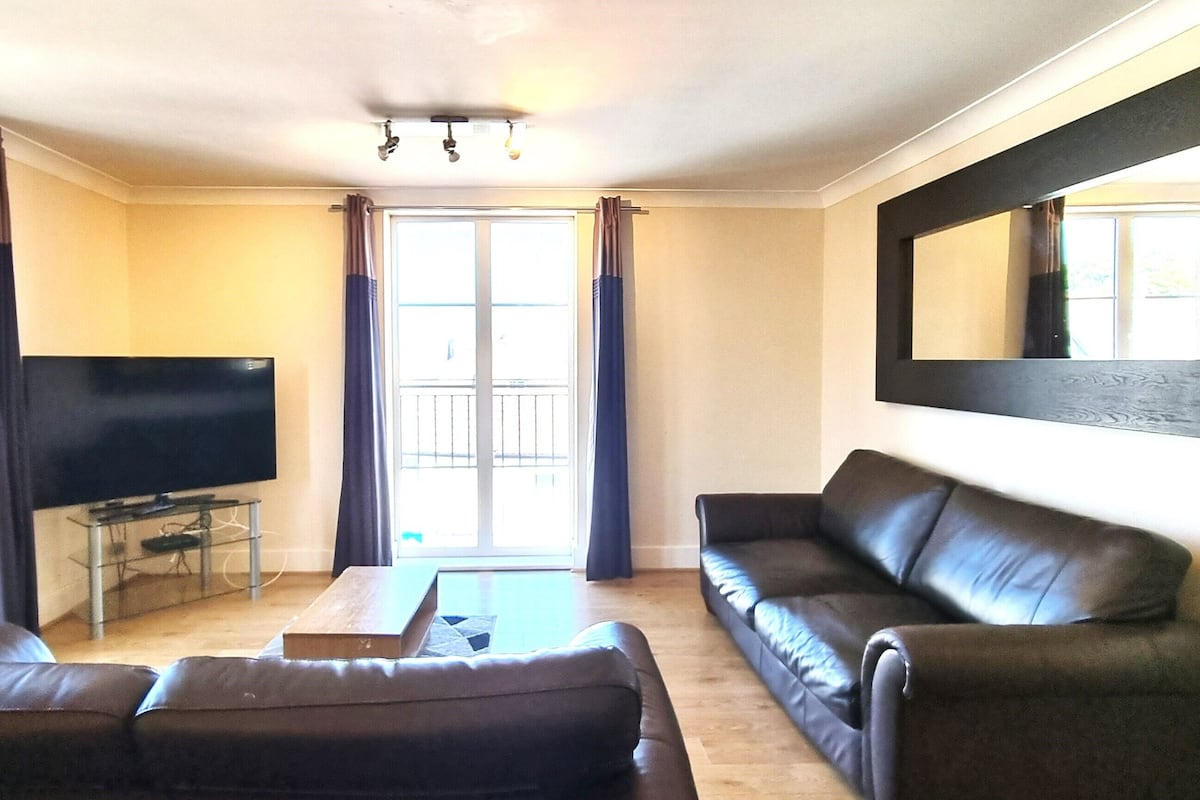 A living room with a large flat-screen TV, leather sofas, a coffee table, and a sliding glass door leading to a balcony.