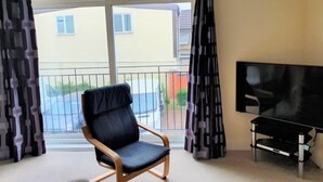 Une pièce avec une grande fenêtre, une chaise noire, un téléviseur posé sur un meuble et un rideau orné d’un motif géométrique.