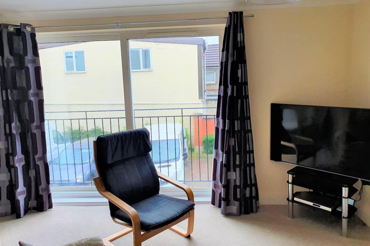 A room with a large window, a black chair, a TV on a stand, and a curtain with a geometric pattern.