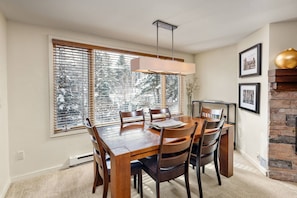 A dining area with a wooden table, chairs, and a pendant light. There is a window with blinds, a fireplace, and framed pictures on the wall.