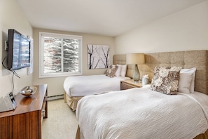 A hotel room with two beds, a TV, a desk with a laptop, and a window with blinds.