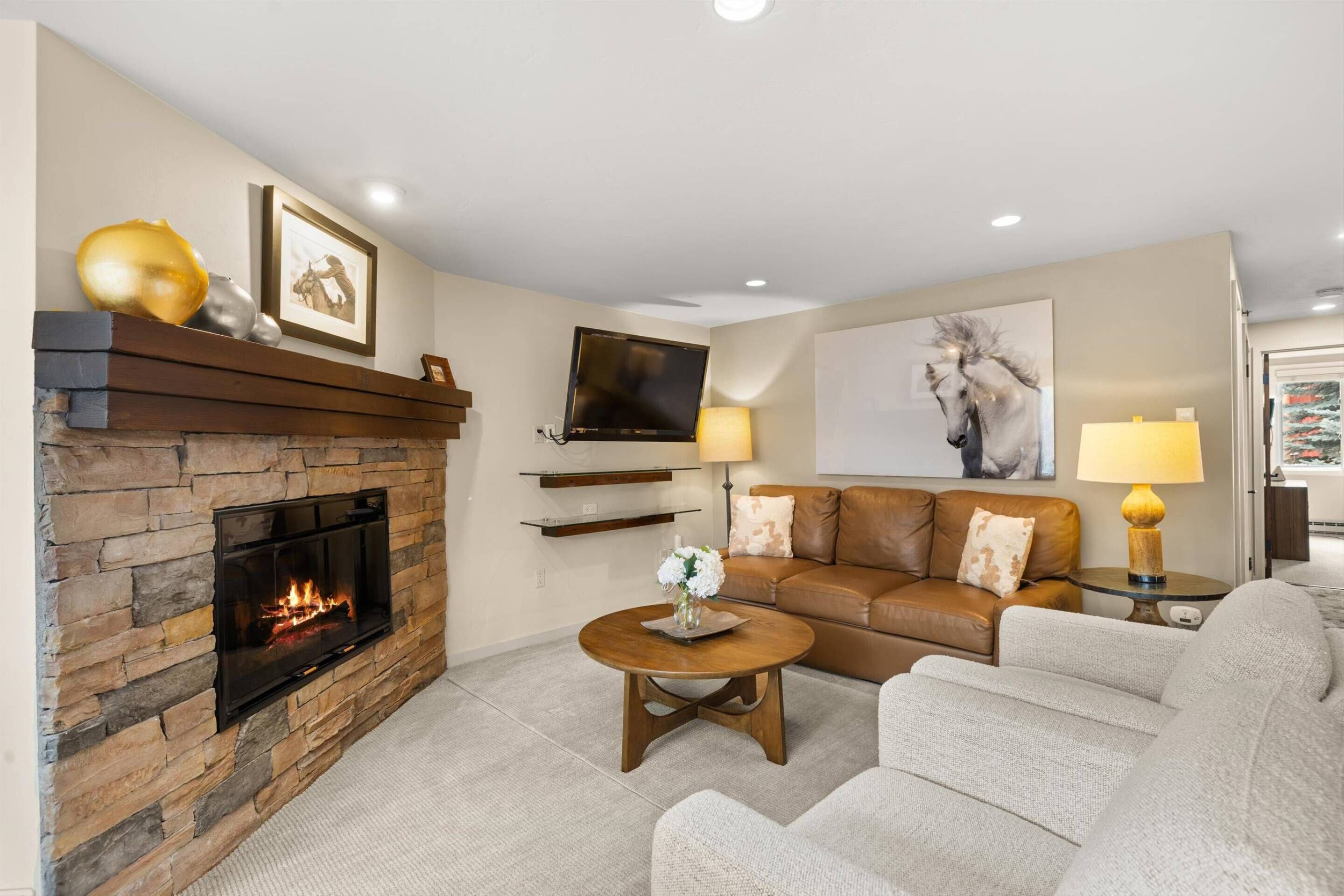 A living room with a stone fireplace, a flat-screen TV mounted on the wall, a brown leather sofa, and a round coffee table with a vase of flowers.
