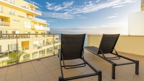 A balcony with two black lounge chairs, a tiled floor, and a view of a yellow building across the water.