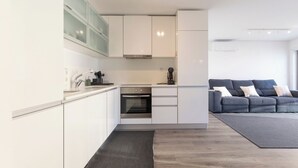 A modern kitchen with white cabinets and a built-in oven, a grey sofa with cushions, and a grey rug.