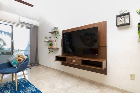 Una sala de estar con un televisor de pantalla plana grande montado en un mueble de madera, un sillón azul y una mesita de noche con objetos decorativos.
