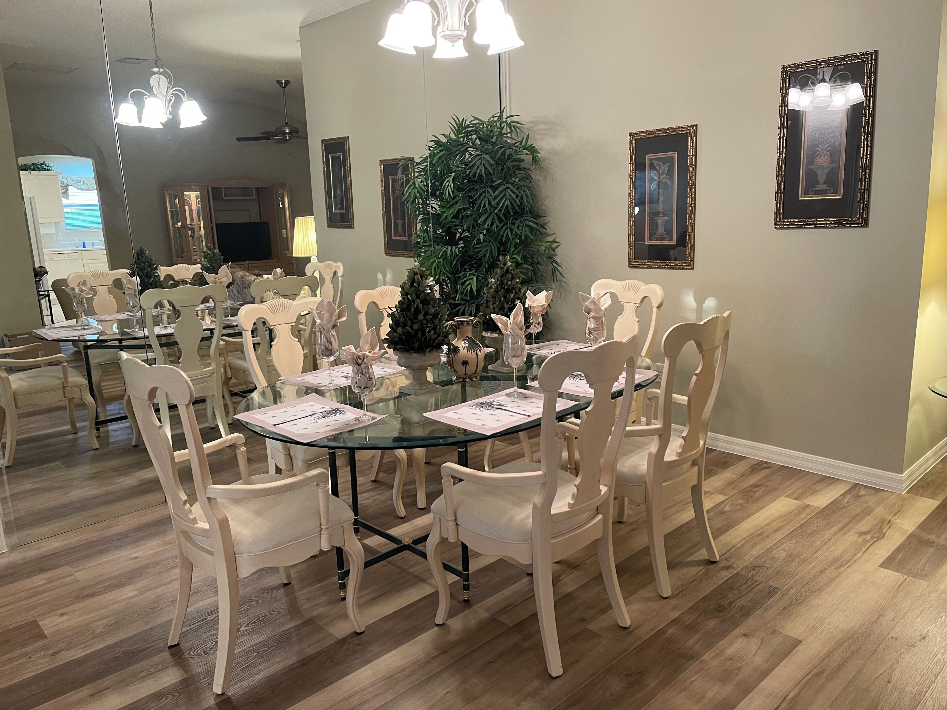 A dining area with a round glass table surrounded by white chairs, a large potted plant, and framed artwork on the walls.