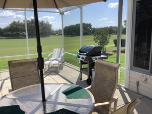 Een overdekt terras met een tafel, stoelen, een barbecue en uitzicht op een groen gazon en bomen.