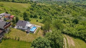 An aerial view of a residential area with a house featuring a swimming pool, surrounded by lush greenery and hilly terrain.