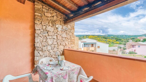 A balcony with a stone wall, a table set with a tablecloth, and a view of houses and greenery in the background. - Beach front apartment in Isola Rossa (Isola Rossa)