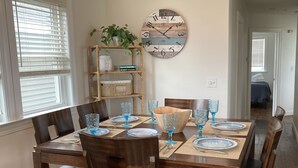 A dining area with a wooden table set for six, surrounded by chairs. A clock is mounted on the wall above the table.
