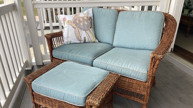A wicker sofa with light blue cushions and a matching ottoman on a wooden deck.
