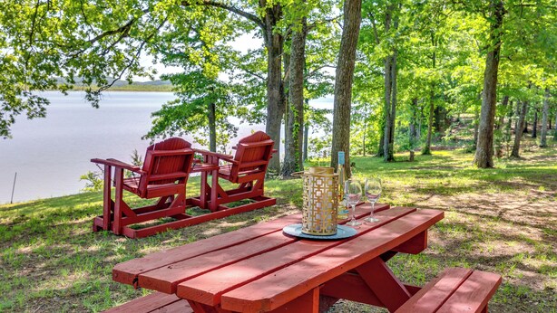 A picnic setup with a red table and chairs, a lantern, and wine glasses on a grassy area near a lake. - Rural Arkansas Vacation Rental w/ Lake Access (Scranton)