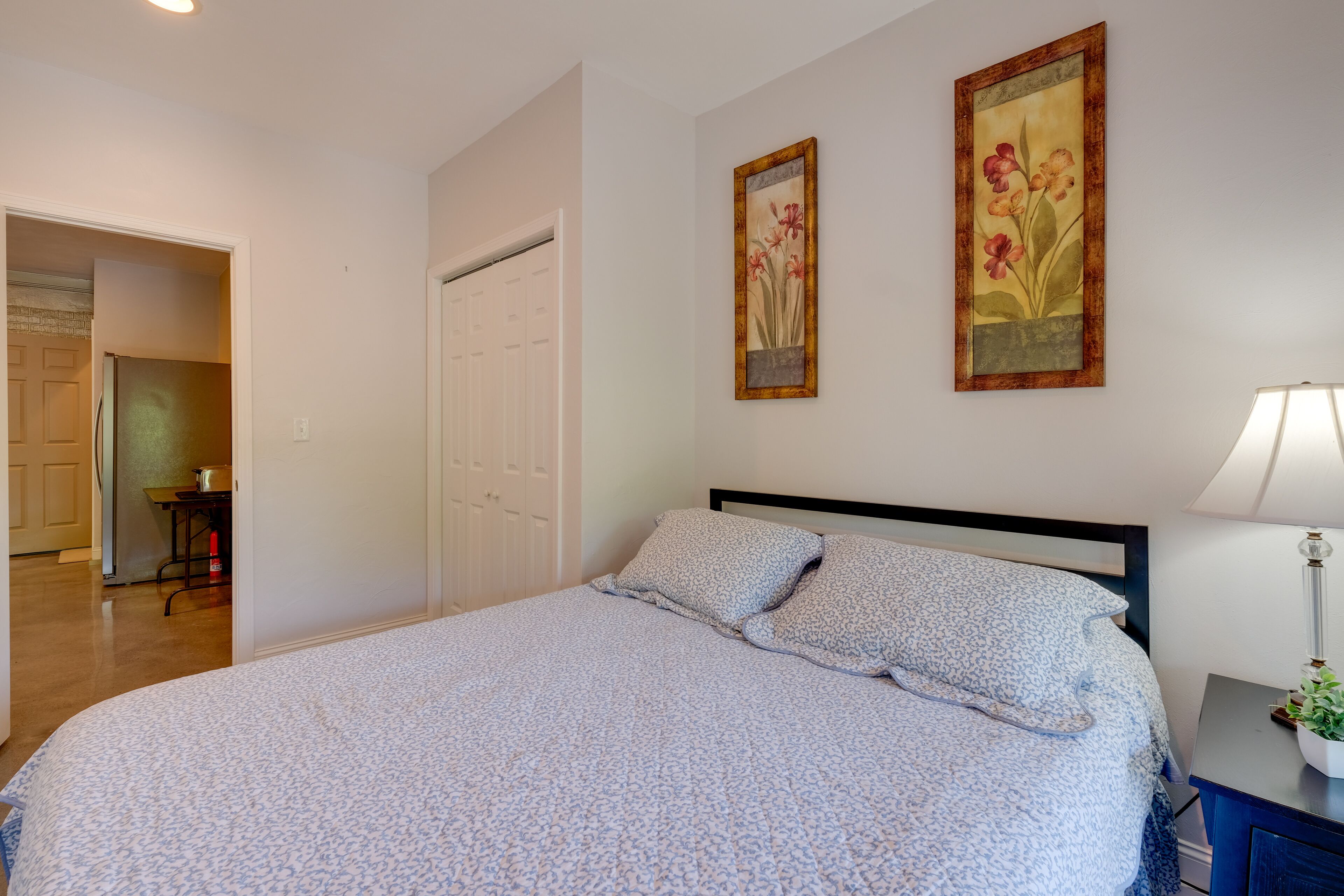 Een slaapkamer met een bed, twee aan de muur hangende schilderijen, een nachtkastje met een lamp, en een deur naar een andere kamer.