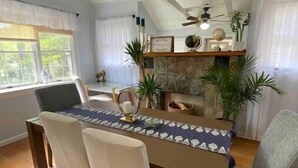 Un comedor con una mesa de madera, sillas blancas, un ventilador de techo y una chimenea de piedra.