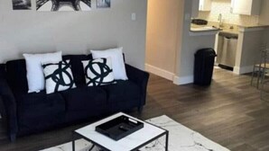 A living room with a black sofa, white cushions, and a coffee table. There are framed pictures on the wall, a kitchen area with a sink and cabinets, and a trash can. - Stylish 2BD/2BA in Arlington | Convenient & Clean! (Arlington)