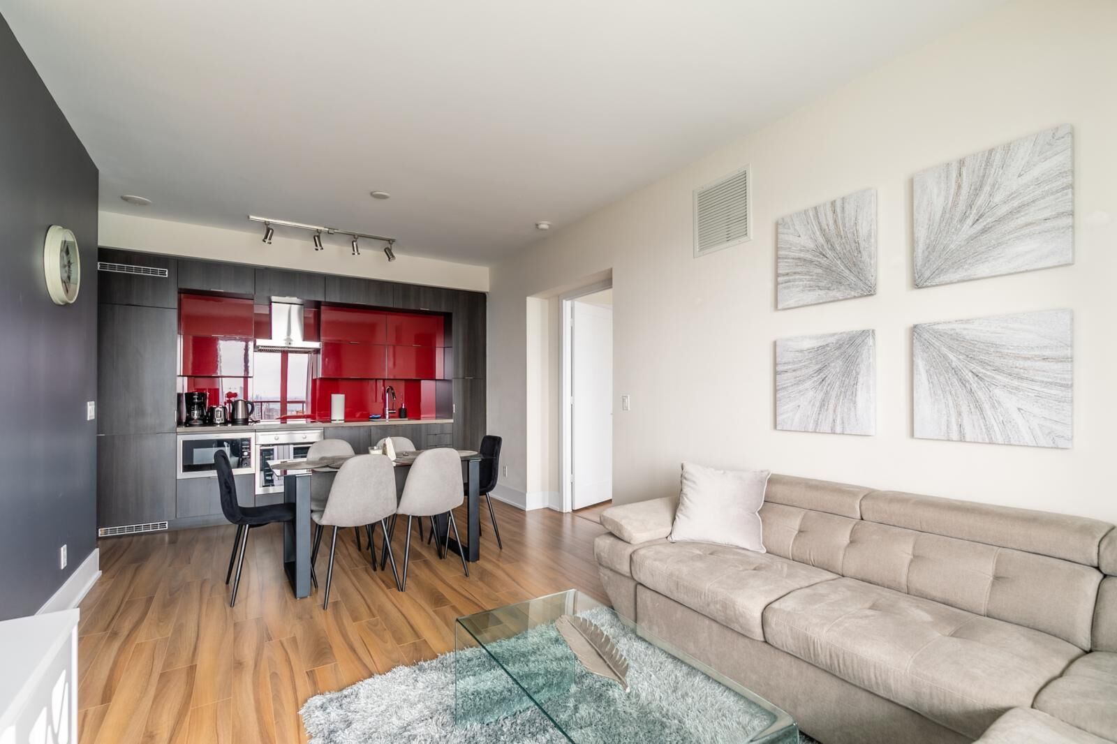A modern living room with a beige sofa, a glass coffee table, and a dining area with a red kitchen in the background.