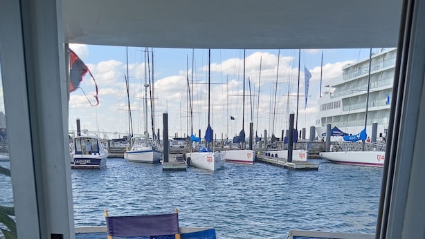 Vue depuis une pièce dotée d’une grande fenêtre donnant sur une marina où sont amarrés plusieurs bateaux. On y trouve deux fauteuils en osier et une petite table sur laquelle repose un panier tressé.