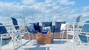 Une terrasse ensoleillée, dotée de chaises longues et d’un panier en osier, qui offre une vue sur une marina remplie de bateaux.