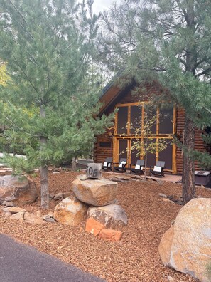 A log cabin with a deck, surrounded by large rocks and pine trees.