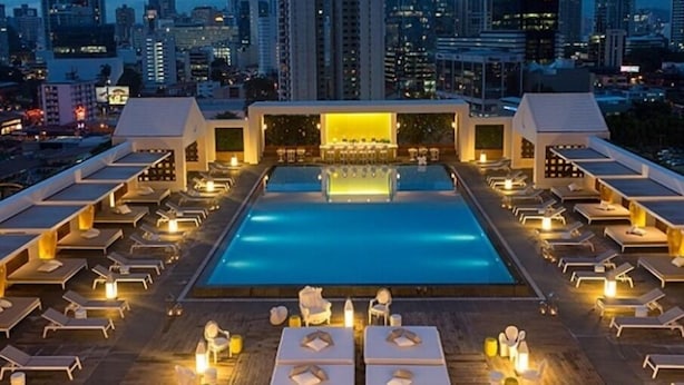 Uma área de piscina no terraço com espreguiçadeiras e mesas, emoldurada por um horizonte urbano ao entardecer.