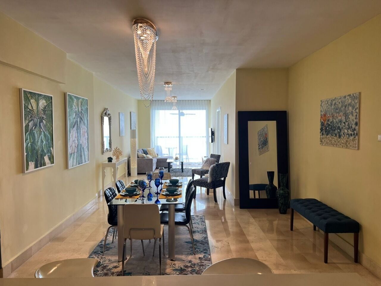Amplio espacio de sala con un comedor listo para cuatro personas, rodeado de asientos cómodos. Un gran candelabro cuelga del techo, y obras de arte decoran las paredes.