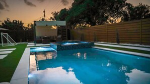 Une piscine entourée d’une bordure en pierre et dotée d’un petit espace avec un jacuzzi, le tout délimité par une clôture en bois et bordé d’une pelouse.