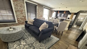Ein modernes Wohnzimmer mit einem dunkelblauen Sofa, einem gemusterten Teppich und einem Hocker. Im Hintergrund ist ein Essbereich mit einem Holztisch und Stühlen zu sehen.