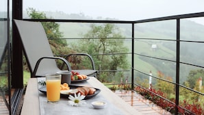 A breakfast table with a glass of orange juice, a plate of toast, and a bowl of soup on a balcony with a view of green hills.