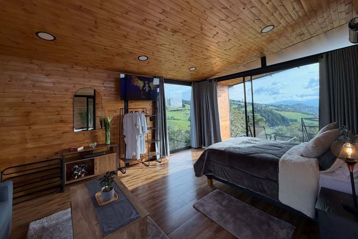 Ein Schlafzimmer mit einem großen Bett, Holzbalkendecke, einem Fernseher, Blick auf die Landschaft und einem Fenster mit Vorhängen.