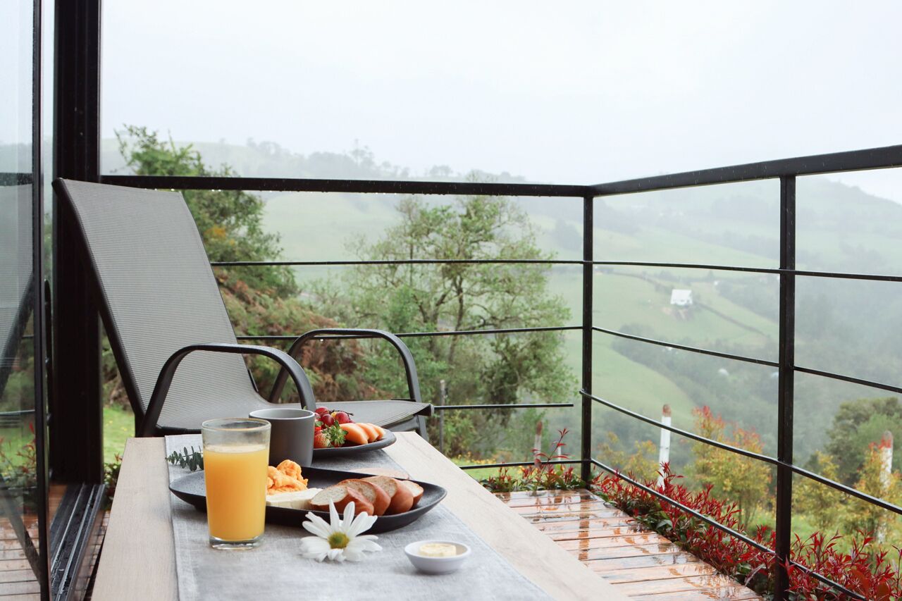 Una mesa de desayuno con un vaso de zumo de naranja, una tostada y un bol de fresas en un balcón con vistas a colinas verdes.