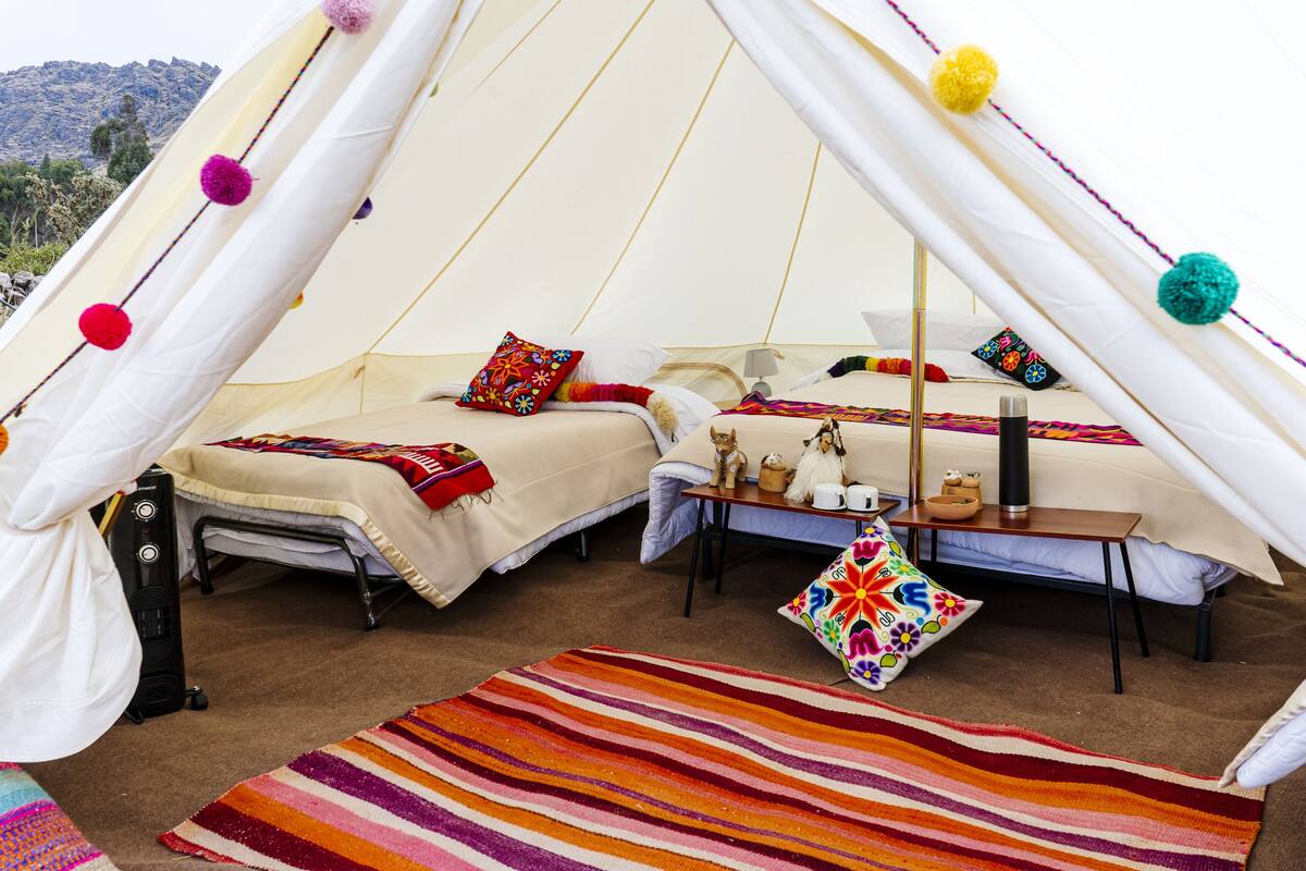 A cozy tented accommodation with two beds, colorful pillows, a striped rug, and a small table with a lamp.