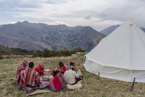 Un grupo de personas sentadas sobre una manta al aire libre, con una tienda de campaña y montañas al fondo.