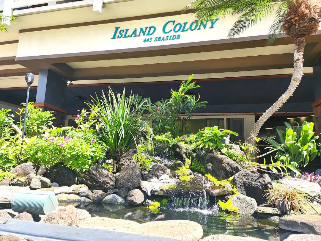 A hotel named 'Island Colony' with a landscaped garden featuring a small waterfall, rocks, and various plants.