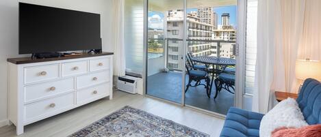 Ein Wohnzimmer mit Flachbildfernseher, einer weißen Kommode, einem gemusterten Teppich, einem blauen Sofa und einem Balkon mit Tisch und Stühlen.