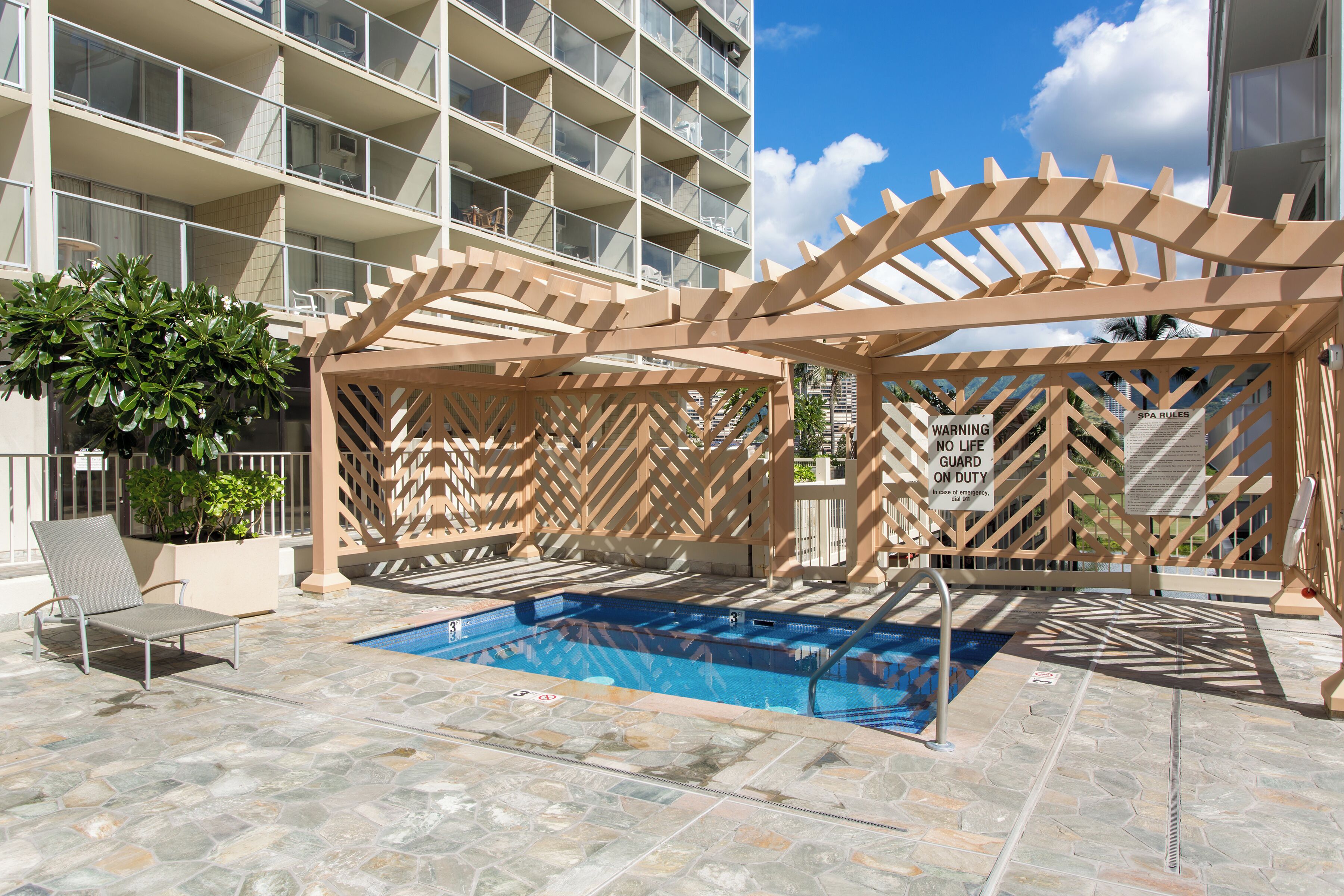 Ein Poolbereich mit einer Holzpergola, Liegestühlen und einem Pool mit blauer Fliesenbordüre.