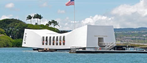 Ein weißes Gebäude mit einem markanten, gewölbten Dach, einer Fahnenstange mit der US-amerikanischen Flagge und Blick auf eine Küste mit Palmen und einer in der Ferne liegenden Stadt.