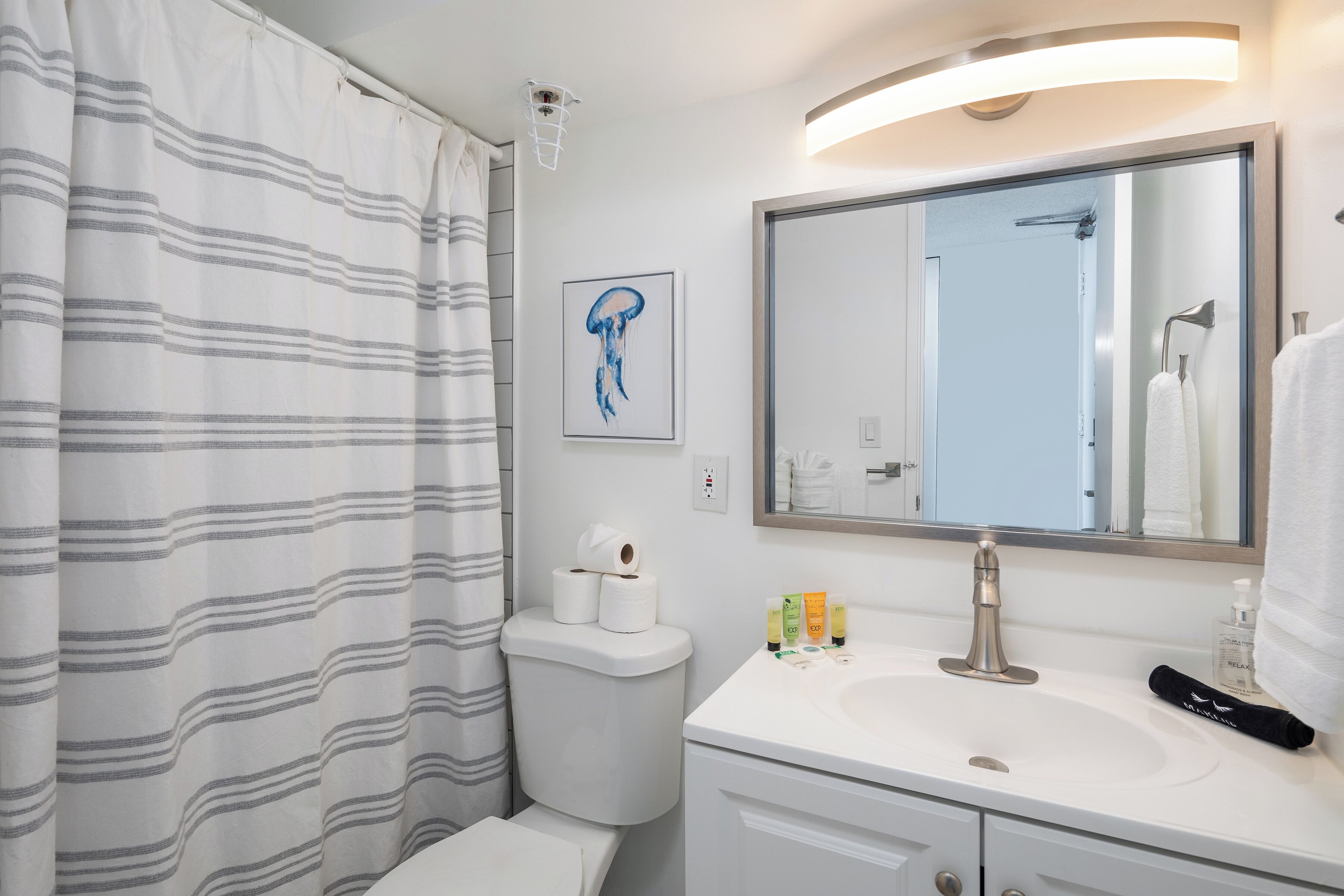 A modern bathroom with a striped shower curtain, a toilet with toilet paper, a sink with toiletries, and a large mirror.