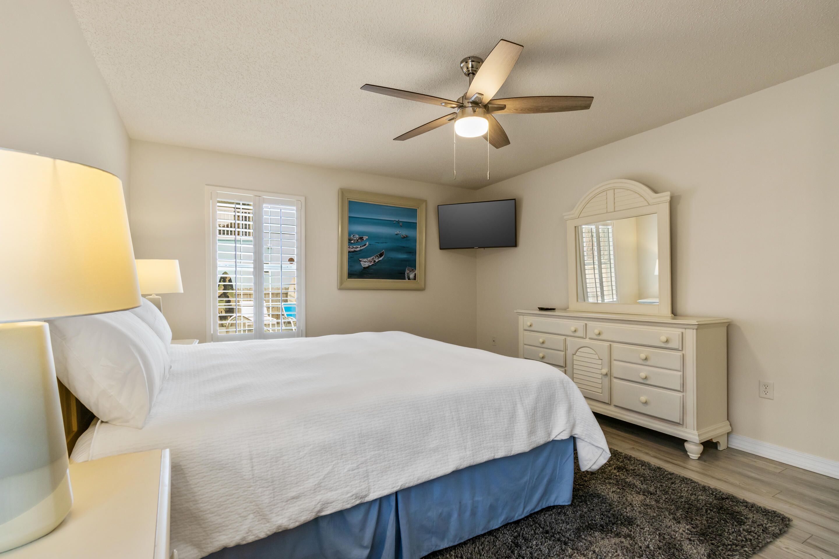 A bedroom with a bed, a ceiling fan, a TV, a dresser, and a window with blinds.